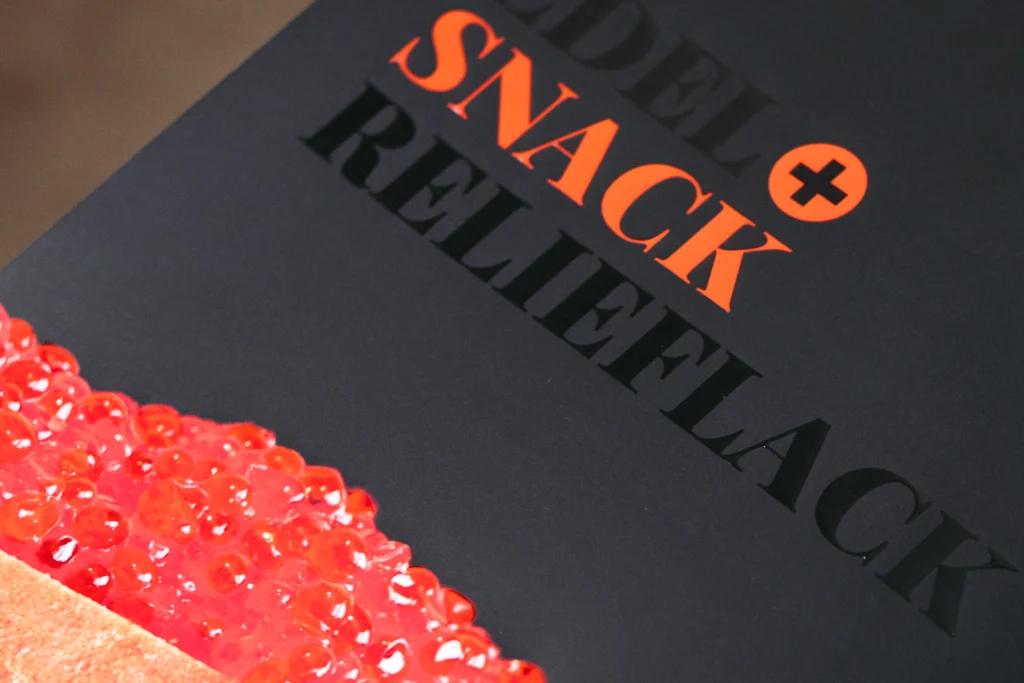 Verpackung mit dem Schriftzug „SNACK RELIEFLACK“ und glänzend lackierten Lachsrogen.