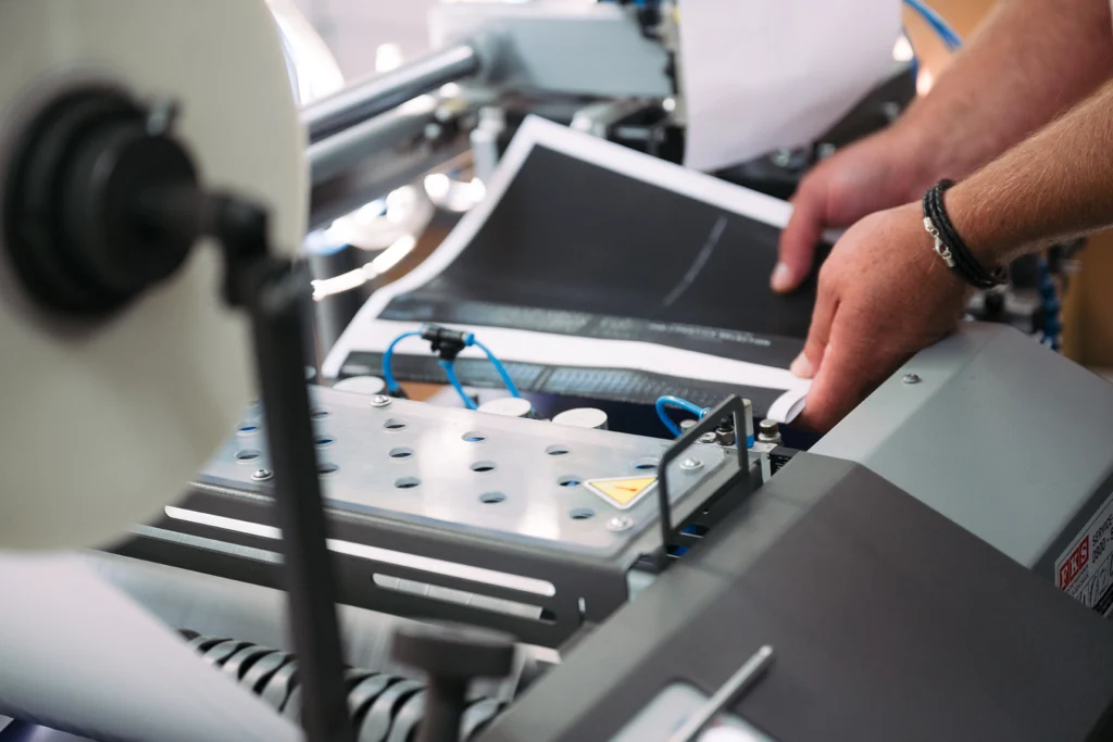 Nahaufnahme einer industriellen Druck- oder Cellophaniermaschine, bei der eine Person bedruckte Seiten manuell einlegt oder überprüft.
