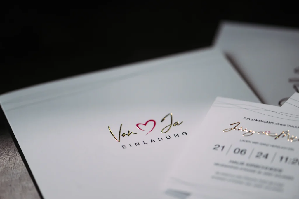 Elegante Hochzeitseinladung mit Glamprint in gold und einem Herzsymbol, das relieflackiert ist.