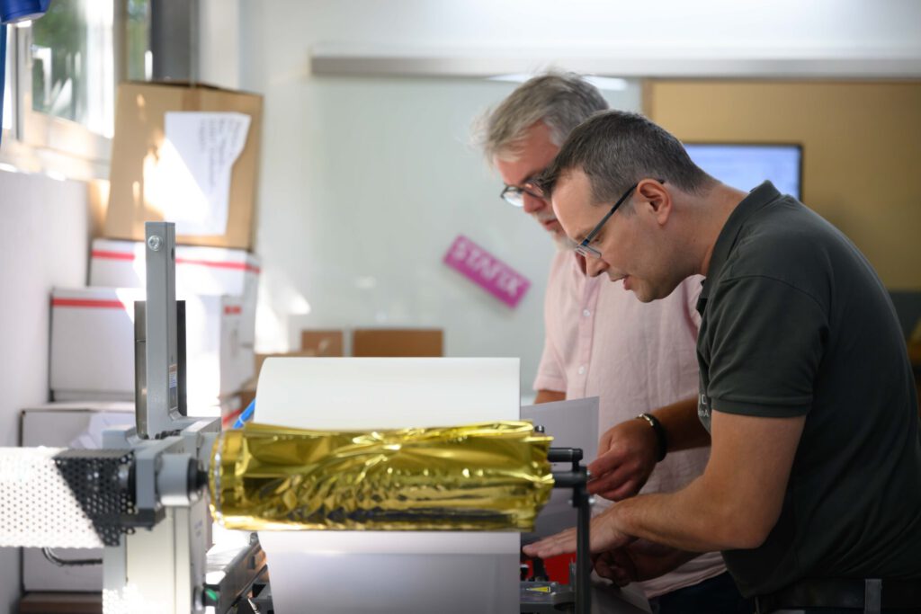 Zwei Mitarbeiter der WARLICH DRUCK RheinAhr sehen sich das Ergebnis der digitalen Heißfolienprägung an.