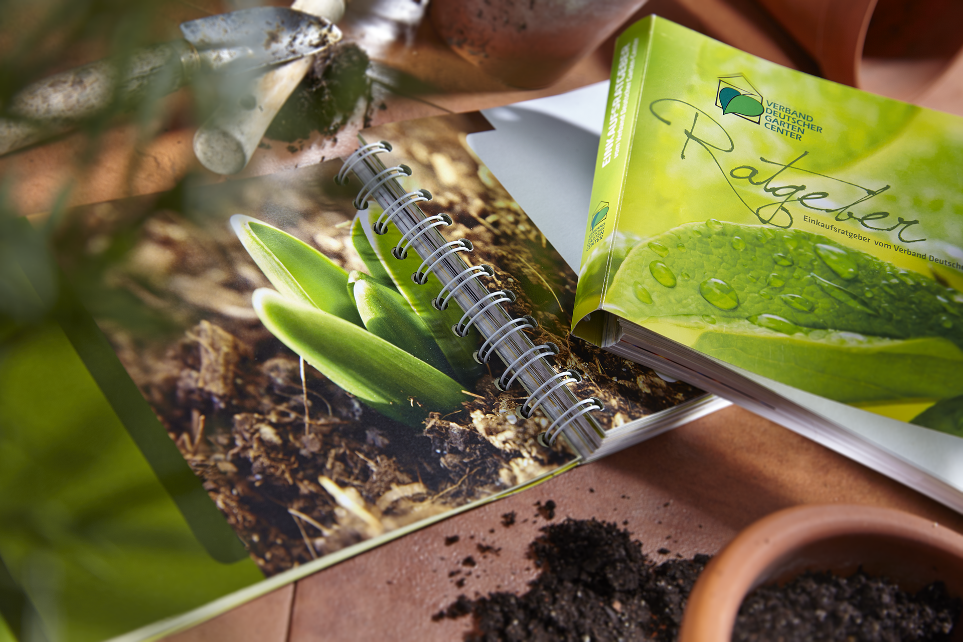 Gedruckter Gartenratgeber mit Spiralbindung und grüner Pflanze, umgeben von Erde und Gartenzubehör
